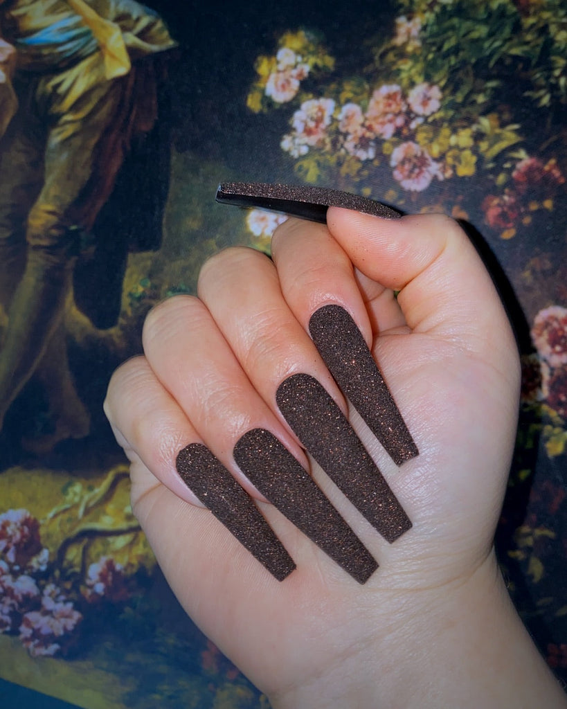 A closed hand displays long, coffin-shaped, brown glitter nails in front of a floral backdrop.
