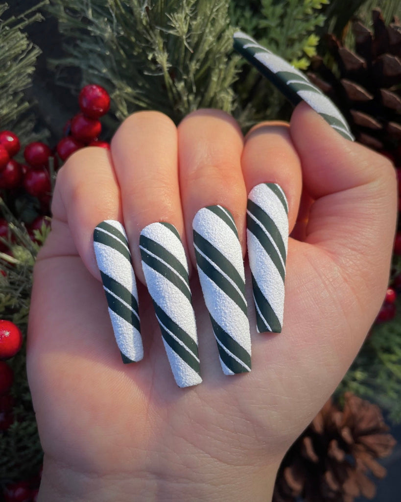 A closed hand displays white, textured nails with bold green stripes cut by fine, white stripes in front of a Christmas tree.