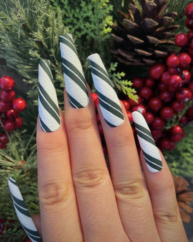Outstretched fingers display white, textured nails with bold green stripes cut by fine, white stripes in front of a Christmas tree.