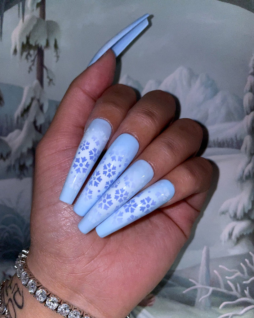 A closed hand displays blue and white ombre nails with airbrushed snowflakes in front of a winter scene.