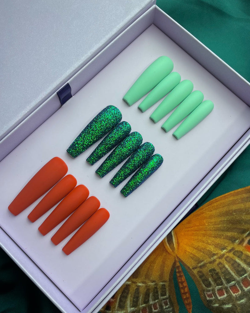 An open white box holds 3 sets of acrylic press-on nails. One light green, one dark green glitter, and one red.