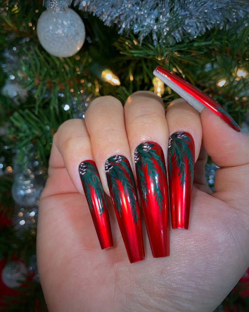 A closed hand displays long, red satin colored nails with hand painted pinecones and spruce leaves in front of a Christmas tree.