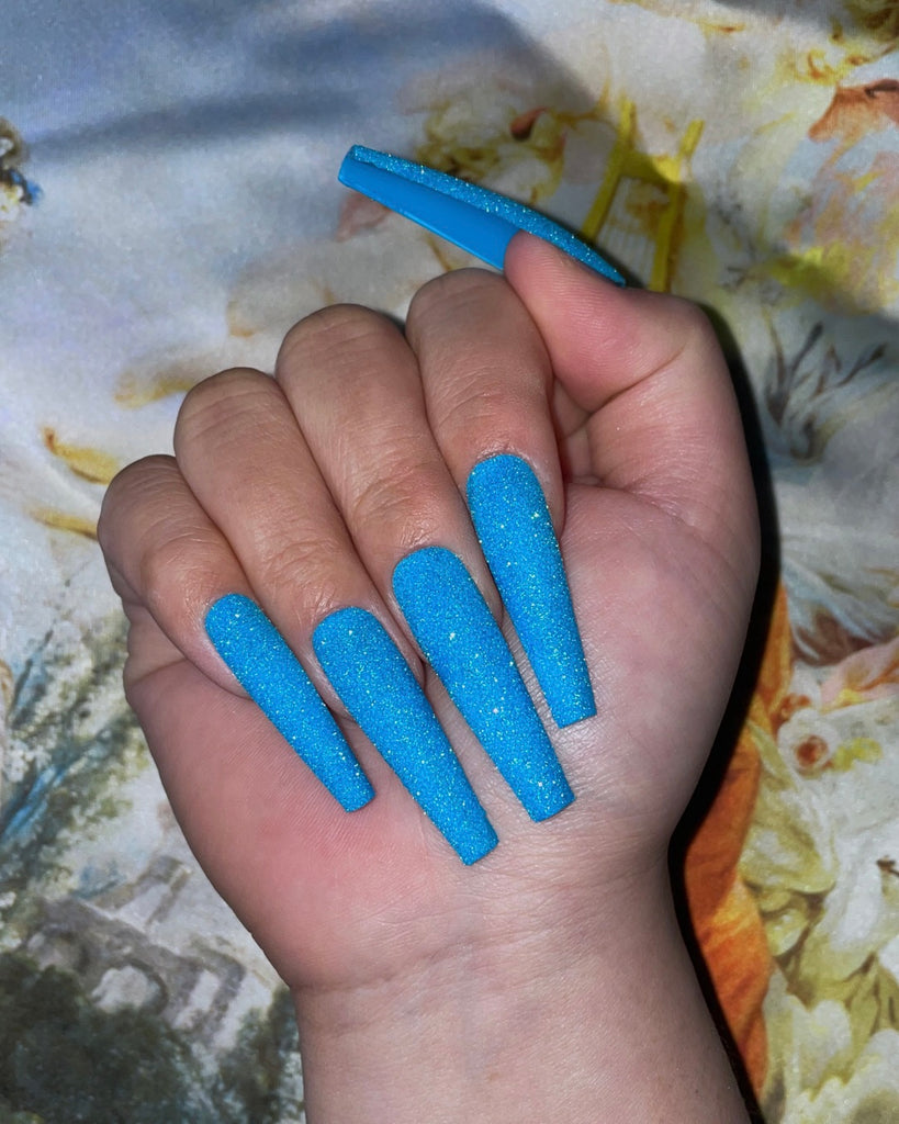 A closed hand displaying long, tapered coffin shaped nails covered with textured light blue glitter in front of a pastel floral background
