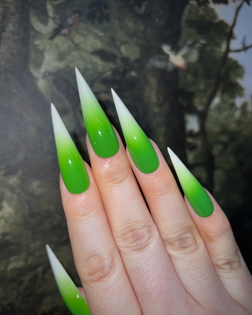 An open hand, palm down displays long pointed nails with a bright green to white ombre in front of an illustrated tree background