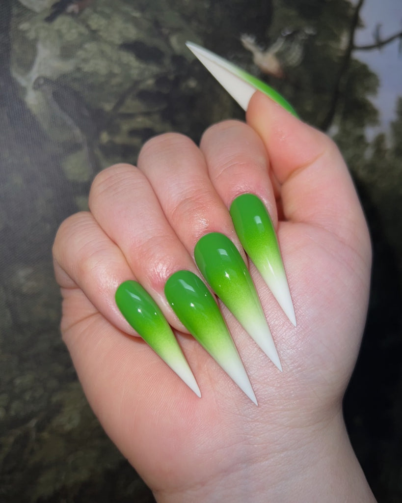 A closed hand displays long, pointed nails decorated with a bright green to white ombre in front of an illustrated tree background